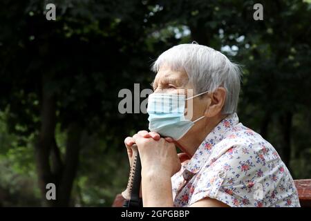 Femme âgée dans un masque de protection assise avec une canne à pied sur un banc dans le parc. Sécurité pendant la pandémie du coronavirus, la vie à la retraite Banque D'Images