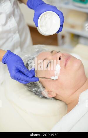 La jeune femme cosmetologiste applique un produit cosmétique sur le visage de la patiente. Spa, procédure cosmétique Banque D'Images