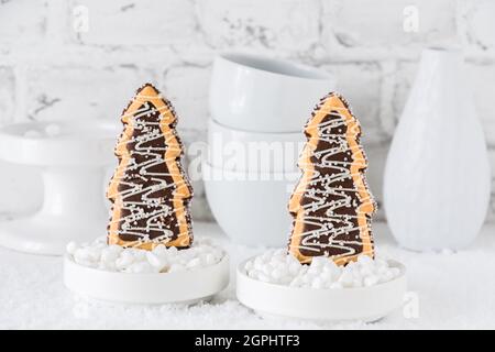 Deux biscuits en forme d'arbres de noël avec chocolat et sucre glacer dans des bols avec du sucre grossier sur fond blanc avec de la vaisselle blanche Banque D'Images