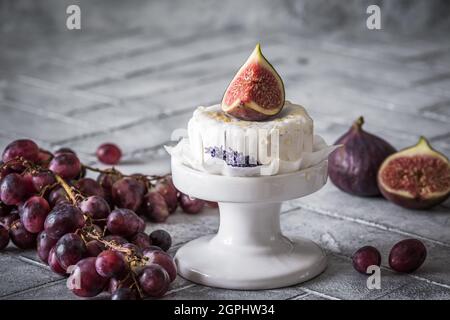 Fromage doux rond entier avec miel et figues sur carreaux gris, décoré de raisins rouges foncés Banque D'Images