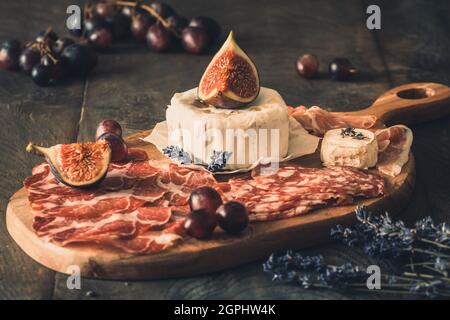 Diverses spécialités de saucisses fumées et un fromage doux sur une planche de bois, décorés de figues et de raisins Banque D'Images