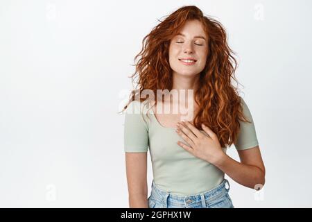 Jeune fille souriante avec des cheveux rouges pensant à quelque chose avec des yeux fermés, tenant la main sur le coeur, rêverie, image scène réconfortant, blanc Banque D'Images