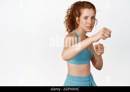 Bonne forme et bonne santé redhead sportswoman faire le poing, l'entraînement en salle de gym, l'entraînement de boxe en vêtements d'activité, debout sur fond blanc Banque D'Images