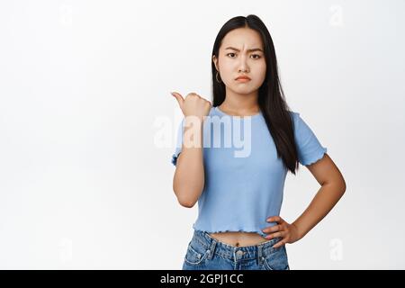 Une jeune femme asiatique en colère pointant du doigt vers la gauche, se moquant et se frognant déçue, se plaignant, mécontente de smth, debout sur fond blanc Banque D'Images