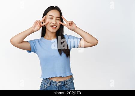 Bonne fille asiatique en forme de clin d'œil, montrant des signes en V kawaii geste près des yeux, vous saluant, debout dans une chemise sur fond blanc Banque D'Images