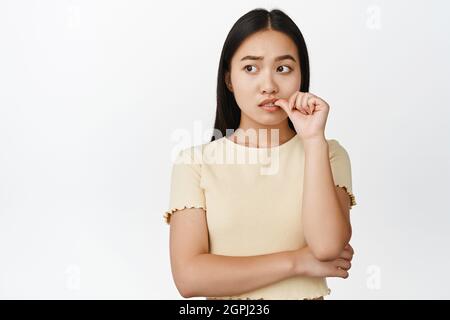 Une fille asiatique nerveuse piquant le doigt et regardant de côté, pensant et se sentent inquiets, debout dans un t-shirt jaune sur fond blanc Banque D'Images