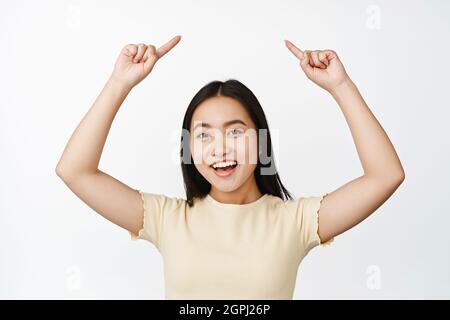 Tour de taille vers le haut de la fille asiatique souriante, pointant les doigts vers le haut, montrant la publicité, debout dans un t-shirt jaune sur fond blanc Banque D'Images