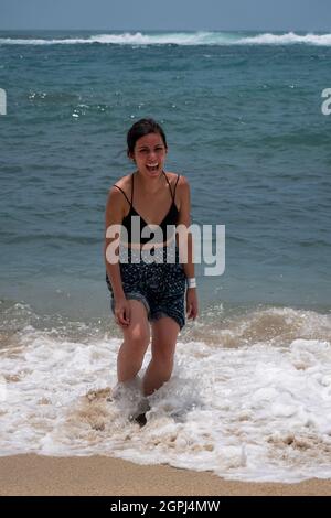 Jeune Latina tenant une paire de sandales et marchant le long de la mer riant beaucoup Banque D'Images