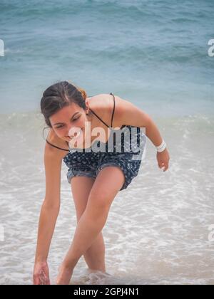 Jeune Latina tenant une paire de sandales et marchant le long de la mer riant beaucoup Banque D'Images