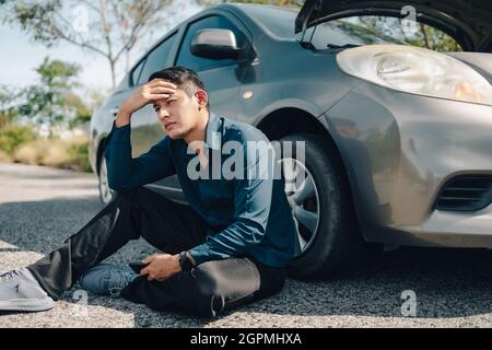 Homme sérieux mal de tête assis à côté d'une voiture en panne sur la route. Concept de voiture brisée. Banque D'Images