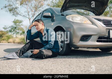 Homme sérieux mal de tête assis à côté d'une voiture en panne sur la route. Concept de voiture brisée. Banque D'Images