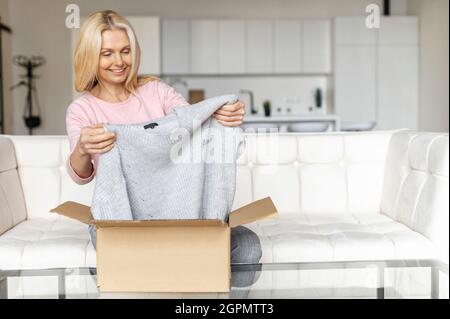 Une femme blonde élégante et satisfaite ouvre une boîte de carton, déballe cadeau reçu, se sentant curieux de l'article commandé dans un magasin en ligne, une cliente satisfaite de la livraison et de l'expédition rapides Banque D'Images