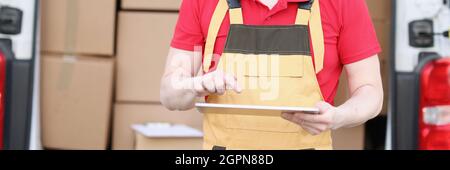 Le joli postier caucasien en costume et casquette vérifie les boîtes aux lettres avec un appareil de tablette dans ses mains Banque D'Images