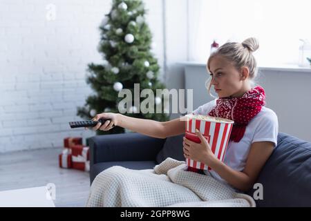 femme malade avec un seau de pop-corn en cliquant sur les chaînes tout en regardant la télévision près d'un arbre de noël flou Banque D'Images