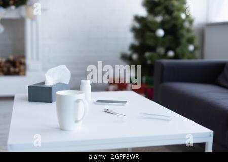 table avec serviettes en papier, contenant de pilules, thermomètre électronique et smartphone près de l'arbre de noël flou Banque D'Images