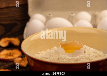 Un œuf de poulet blanc cassé dans un bol avec de la farine de blé, des chips de pomme séchées et plusieurs œufs entiers dans un plateau en carton blanc, gros plan sélectif Banque D'Images