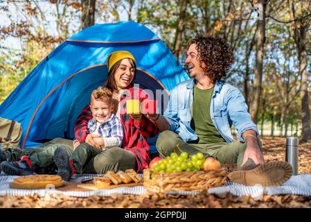 jeune famille heureux campant à l'extérieur avec tente dans la nature d'automne. maman, papa et petit enfant ayant un pique-nique dans la nature. les gens faisant un toast Banque D'Images