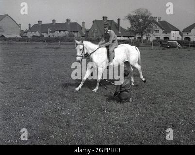 Années 1950, historique, à l'extérieur d'un champ lors d'un événement, une jeune fille pilote sur un cheval sautant au-dessus d'un poteau en bois tenu par deux jeunes garçons, West Sussex, Angleterre, Royaume-Uni. Banque D'Images