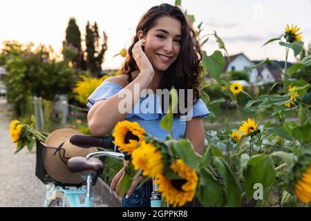 Belle jeune femme marchant dans un lotissement tout en poussant son vélo turquoise avec des tournesols dans un panier. Sunbeam en arrière-plan. Banque D'Images