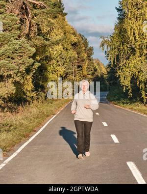 Femme active senior avec cheveux gris courant le long de la route pavée de forêt le jour ensoleillé d'automne. Banque D'Images