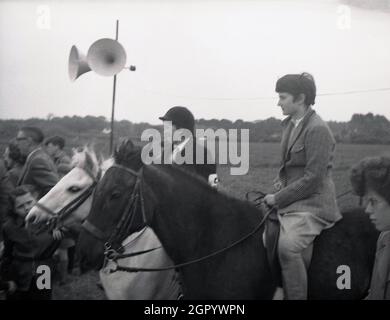 Années 1950, historique, deux jeunes sur leurs chevaux, l'un portant un casque, l'autre pas, debout à côté de spectateurs regardant une compétition équestre, West Sussex, Angleterre, Royaume-Uni. Banque D'Images