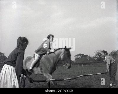 Années 1950, historique, à l'extérieur d'un champ à un événement équestre, une jeune fille pilote sur un cheval sur le point de sauter sur un poteau en bois hend par deux jeunes filles, West Sussex, Angleterre, Royaume-Uni.Les cavaliers sur des chevaux sautant sur des obstacles ou des barrières, parfois tenus à la main comme vu ici, sont une vue commune à des spectacles équestres, avec le saut de spectacle faisant partie d'un groupe d'équitation anglaise des événements équestres. Banque D'Images