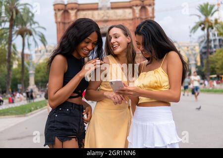 Groupe de femmes millénaires multiraciales heureuses dans des tenues élégantes utilisant un téléphone mobile et en choisissant un itinéraire touristique lors de la visite de Barcelone pendant les vacances d'été Banque D'Images