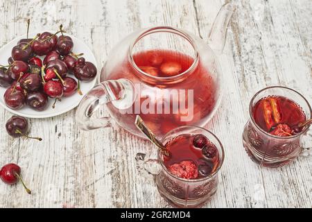 Thé aux fruits dans une théière en verre avec une assiette de cerises rouges et deux tasses de thé sur une table en bois Banque D'Images