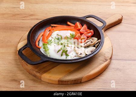 Petit-déjeuner anglais dans une poêle avec œufs frits, poivre, champignons et tomates sur un support en bois Banque D'Images