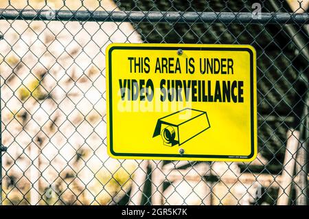 Panneau de surveillance vidéo jaune boulonné sur une clôture à maillons de chaîne avec un lettrage noir lisant CETTE ZONE EST SOUS SURVEILLANCE VIDÉO. Banque D'Images