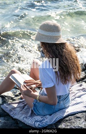 Jeune femme méconnaissable assise près de la mer lisant un livre Banque D'Images