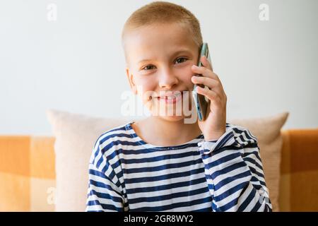 Un garçon blond émotionnel parle au téléphone. Assis sur un canapé dans des vêtements décontractés souriants. Discuter avec vos amis et votre famille. Bonne enfance Banque D'Images