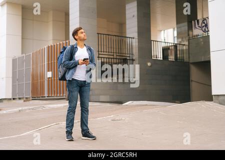 Vue avant d'un beau messager masculin confus avec sac à dos thermo à l'aide de l'application de navigation sur le téléphone situé dans la rue de la ville. Banque D'Images