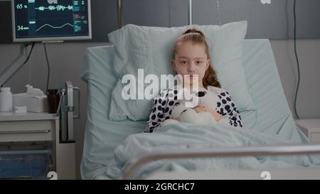 Portrait d'un petit enfant solitaire regardant dans l'appareil photo tenant un ours en dents dans les mains se reposant dans le lit souffrant d'infection. Enfant en attente d'un traitement de récupération pendant un examen médical dans la salle d'hôpital Banque D'Images