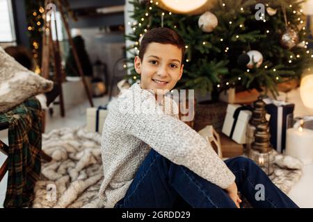 Portrait d'un adolescent élégant et heureux assis près d'un arbre de Noël décoré. Jeune homme regardant directement la caméra, souriant, fêtez le nouvel an Banque D'Images