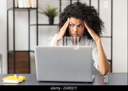 Une femme d'affaires afro-américaine fatiguée dans une chemise décontractée, assise à la réception devant un ordinateur portable, a contrarié une employée de bureau de course mixte se sent mal et a l'épuisement Banque D'Images
