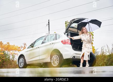 Belle femme sur la route près de sa voiture blanche arrêtée avec pneu de voiture crevé. Femme chauffeur demandant de l'aide, tenant un parapluie, assis sur la roue de secours. Banque D'Images