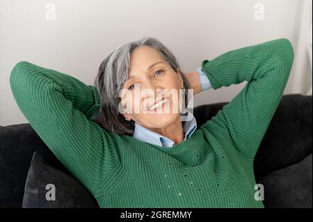 Gros plan d'une femme mûre et souriante se reposant sur le canapé ou le canapé, placé ses mains derrière la tête, se détendant après une longue journée de travail, ayant une grande humeur, et rêvant de jour Banque D'Images