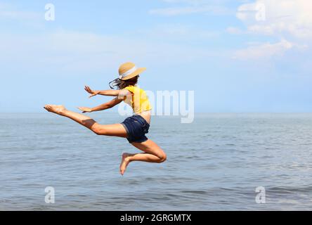 jeune fille skinny avec chapeau de paille et t-shirt jaune effectue un saut en hauteur au bord de la mer en été Banque D'Images