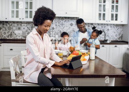 Femme concentrée travaillant sur une tablette numérique dans une cuisine lumineuse pendant que son mari nourrit deux filles en arrière-plan. La famille afro-américaine chez elle. La technologie pour les modes de vie. Banque D'Images