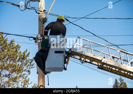 Cliché vertical d'un technicien d'une entreprise d'alimentation électrique, portant un kit de sécurité, assis sur le dessus d'une échelle fixée au poteau électrique Banque D'Images