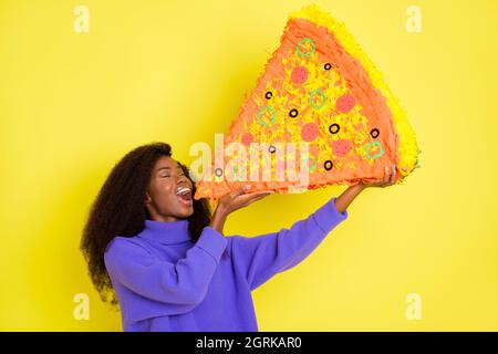 Photo de la peau sombre positive fille yeux fermés les mains tiennent grande pizza bouche ouverte isolée sur fond jaune Banque D'Images