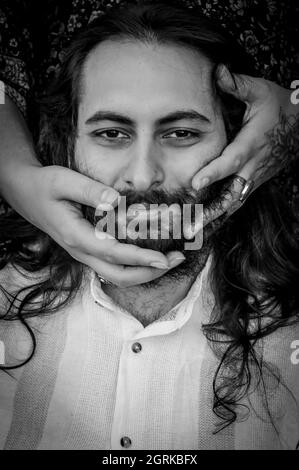 Portrait noir et blanc d'un jeune homme barbu et aux cheveux longs avec les mains d'une femme qui caressent sa barbe Banque D'Images