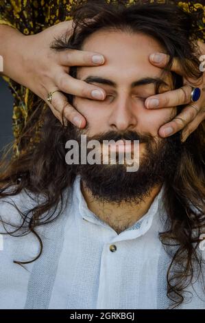 portrait d'un jeune homme barbu et aux cheveux longs avec les mains d'une femme qui caressent son visage et qui couvre ses yeux Banque D'Images