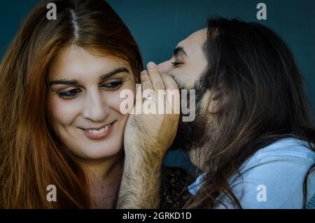 Portrait d'un jeune couple où l'homme barbu avec de longs cheveux raconte un drôle de secret à la femme avec des cheveux rouges et elle sourit Banque D'Images