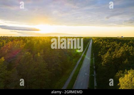 Vue aérienne depuis un drone d'une route en béton qui traverse les forêts d'automne dans des couleurs vert jaune or. Forêt en période dorée et autoroute vide en automne sur un coucher de soleil lumineux. Chaussée entre les arbres Banque D'Images