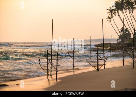 Les pêcheurs traditionnels stilt bâtons pour la pêche dans l'océan wavesat coucher de soleil près de Mirissa, Ahangama, Sri Lanka Banque D'Images