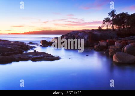 Lever du soleil dans la baie de Binalong des baies de Fire Coast en Tasmanie, en Australie. Rochers recouverts de lichen rouge pittoresque. Banque D'Images