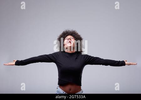 Joyeuse et exubérante jeune Black femme célébrant avec les bras ouverts et sa tête projetée avec un sourire éclatant sur un fond gris studio avec copyspa Banque D'Images