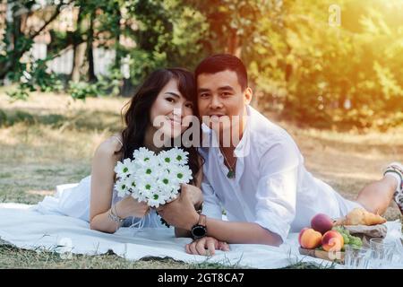 Un couple asiatique charmant s'est happé pique-niquer dans un parc, allongé sur leurs estomacs. Femme tenant un bouquet, se reposant dans des vêtements blancs dans une ombre d'arbre. Banque D'Images